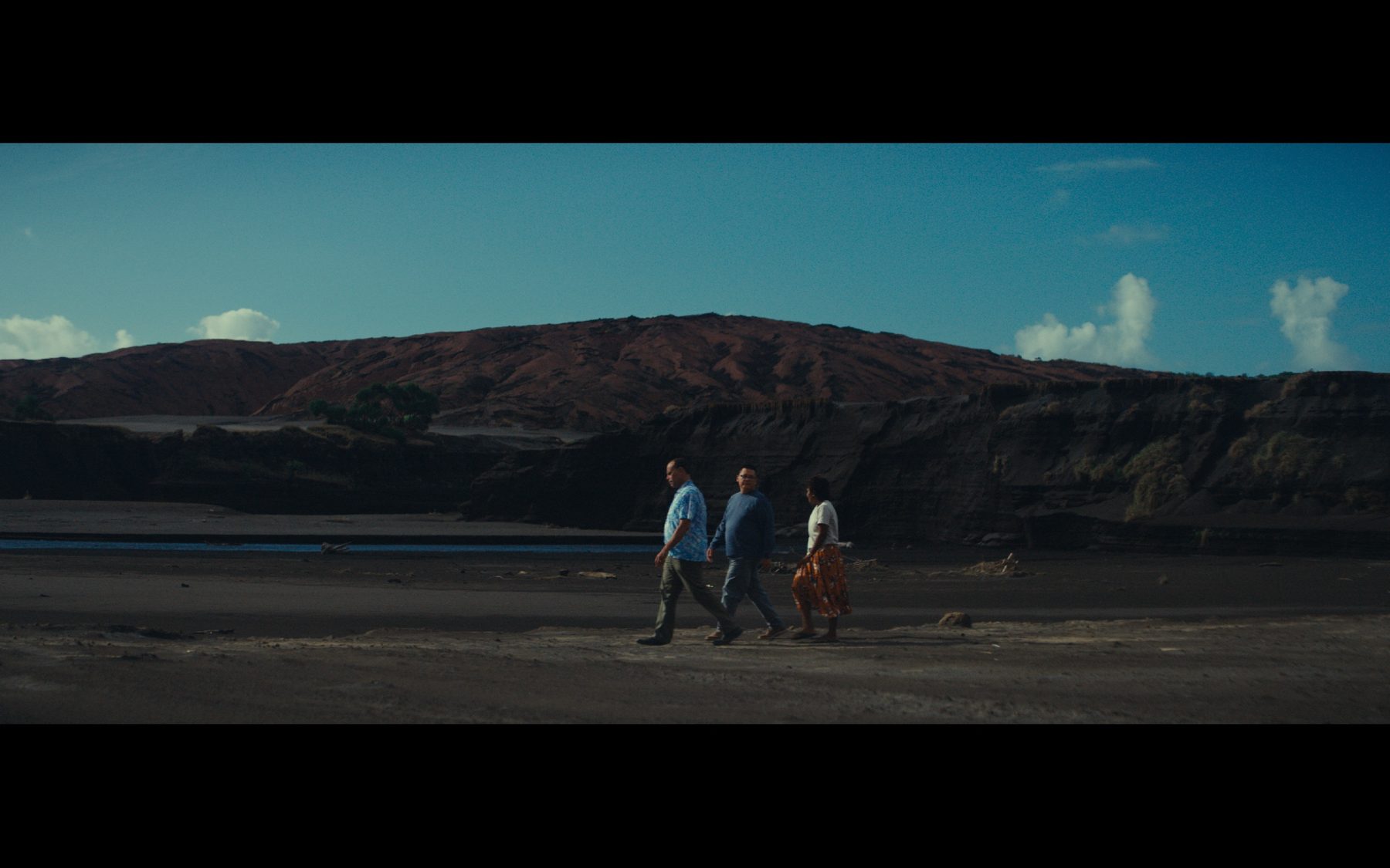 Three people walking a dark shore beneath a red ridge