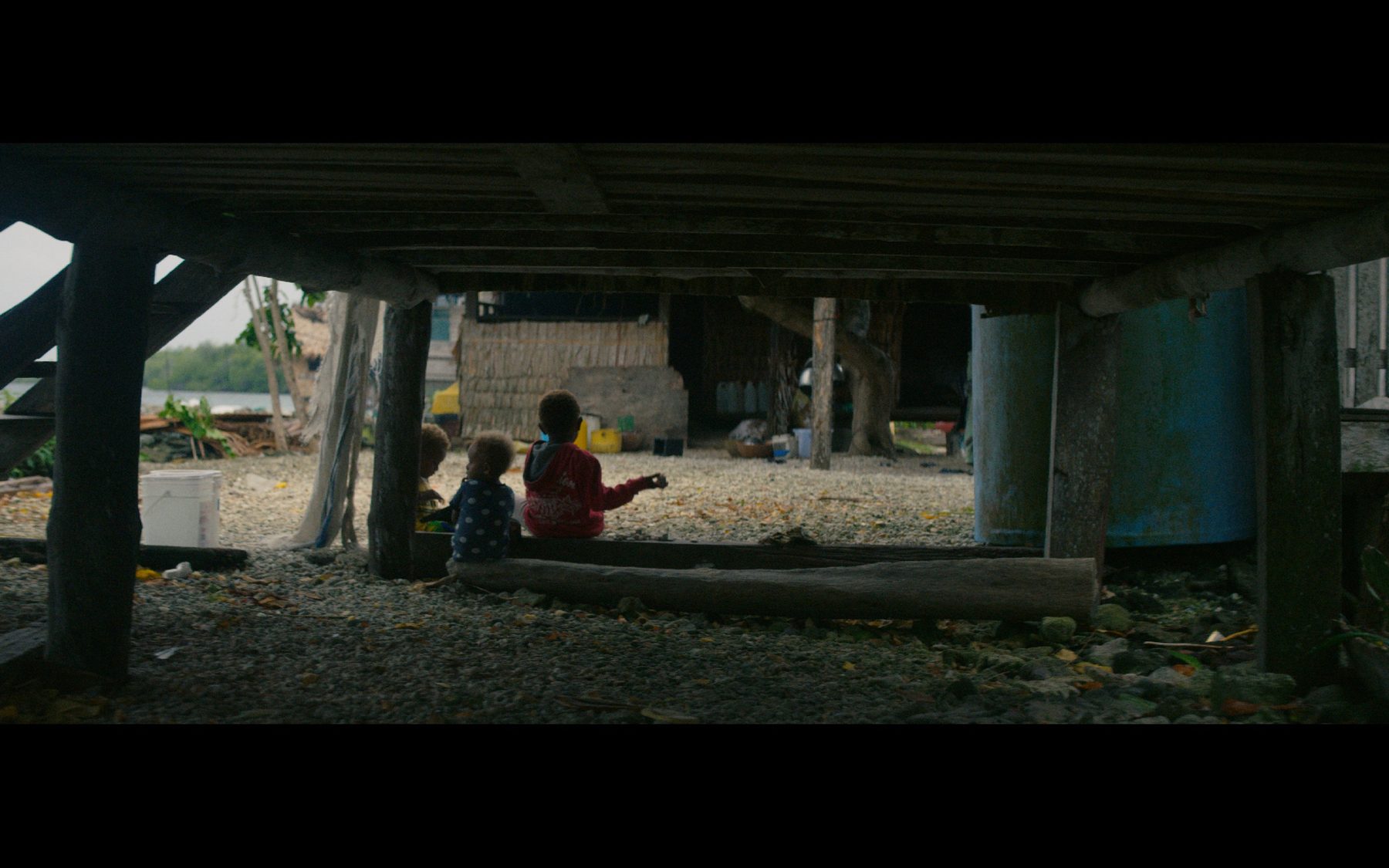 Children sitting under a raised house by the shore